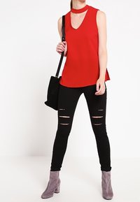 Top rosso senza maniche con scollatura a V e dettaglio choker, abbinato a jeans skinny neri strappati e stivaletti grigi alla caviglia, con una borsa nera.