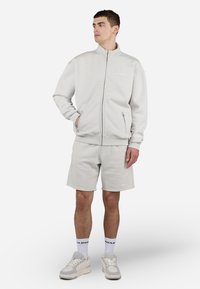 Giacca zip grigia chiara con pantaloni corti coordinati, entrambi realizzati in tessuto morbido. Presenta tasche laterali e un logo discreto sul petto e sui pantaloni.