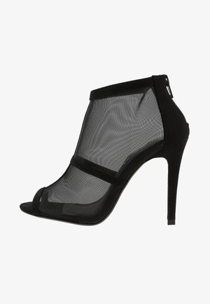 Botte de cheville noire à talons hauts avec panneaux en maille, bout ouvert et fermeture éclair. Présente un talon aiguille fin et des accents en daim.