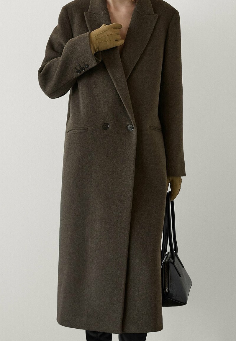 Personne portant un long manteau marron et des gants moutarde tenant un sac à main noir sur un fond uni.