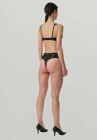 Kvinna som står med ryggen mot kameran iförd svart spetslinne och svarta högklackade skor mot en enfärgad ljus bakgrund.