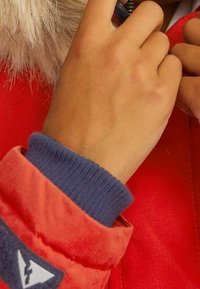 Czerwona kurtka puchowa z granatowym ściągaczem, futrzanym kapturem i szarym zamkiem błyskawicznym. Ręka dostosowuje zamek, ukazując naturalną teksturę skóry.