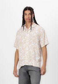 Kortermet, lys beige skjorte med et hvitt abstrakt mønster, knappestolpe foran og en avslappet passform. Kombinert med grå jeans.