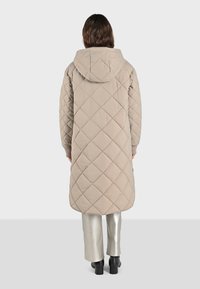 Cappotto lungo beige trapuntato con cappuccio, caratterizzato da motivi a diamante, tasche laterali e polsini a coste. Il modello indossa pantaloni argento e stivali neri.