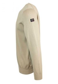 Maglione beige a maglia con maniche lunghe, polsini e orlo a coste. Presenta una toppa con logo sulla spalla sinistra. Texture liscia e design aderente.