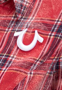 Czerwona koszula bawełniana w kratę z białym haftowanym logo. Tkanina prezentuje kontrastujące granatowe, żółte i czarne linie w wzorze.