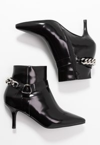 Botines de charol negro con punta afilada, tacones delgados y cremallera lateral. Presentan un detalle de cadena alrededor del tobillo.