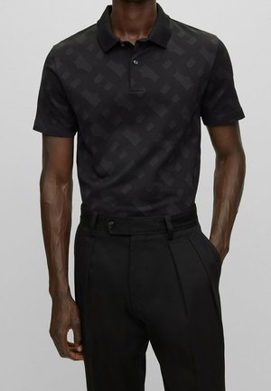 Mann trägt ein schwarzes Polohemd mit kurzem Arm und einem dezenten geometrischen Muster sowie hoch taillierte schwarze Hose mit Falten.