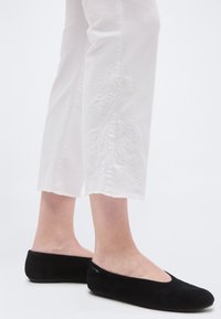 Jambes portant un pantalon blanc court orné de broderies florales et des chaussures plates noires à enfiler sur un fond blanc uni.