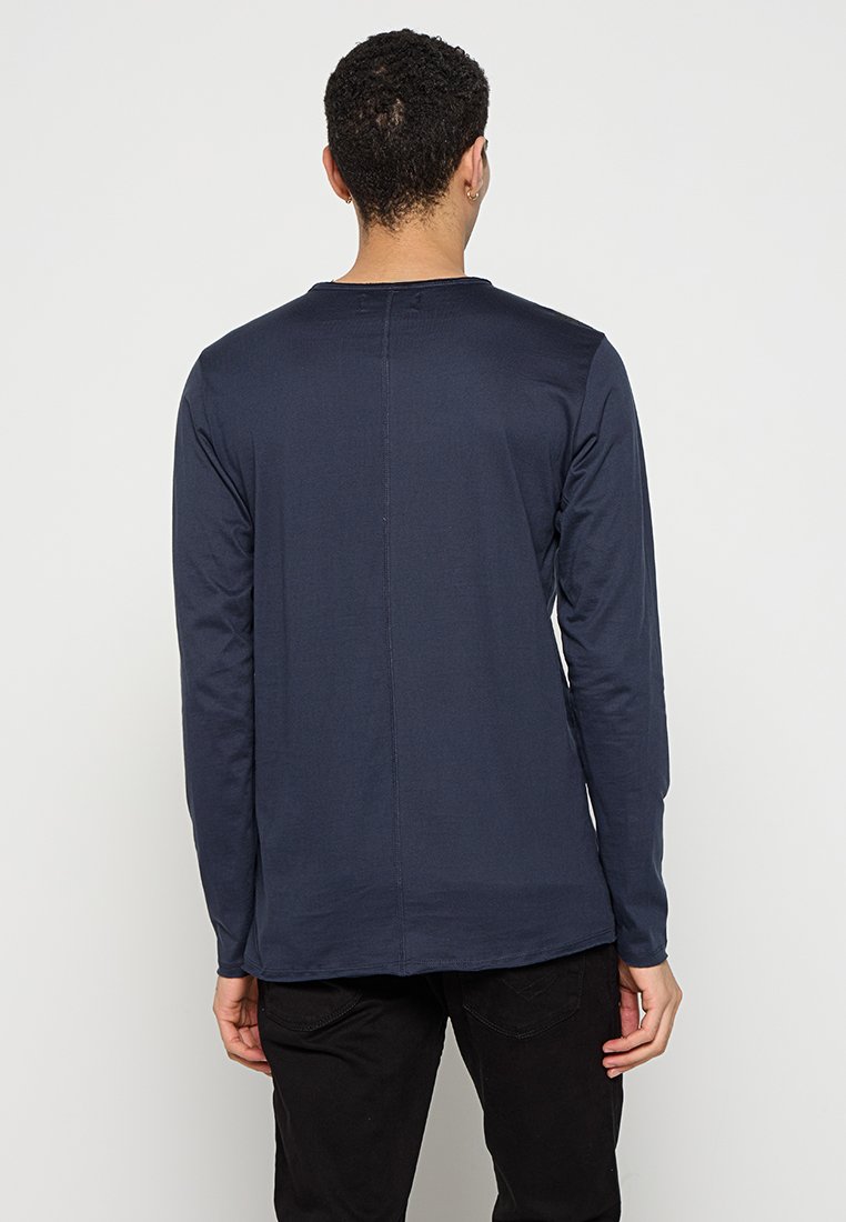 Marineblaues Langarmshirt, weiches Material, Rundhalsausschnitt, schlichtes Design, Nahtdetails am Rücken. Getragen mit schwarzen Hosen, minimalistischer Stil.