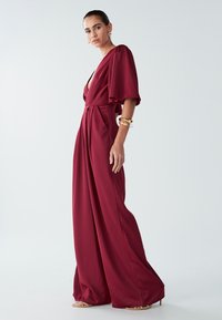 Mujer con un mono fluido de color rojo intenso, de piernas anchas y mangas cortas, que lleva pendientes dorados, múltiples pulseras y sandalias de tacón alto en tono nude.