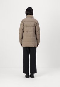 Personne debout de dos portant une doudoune beige, un pantalon large noir et des bottes à plateforme noires sur un fond blanc uni.