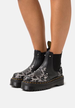 Botines negros y grises con un patrón texturizado, paneles laterales elásticos y una suela gruesa y estriada con detalles de costuras amarillas.