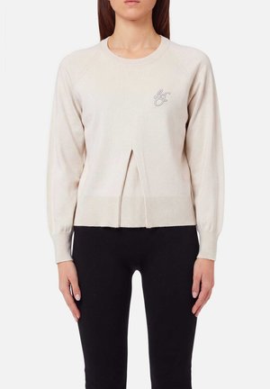 Maglione beige a maniche lunghe con piega frontale e piccole iniziali ricamate, abbinato a pantaloni neri aderenti su una persona in piedi.