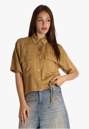 Donna che indossa una camicia senape larga a maniche corte con due tasche sul petto e jeans azzurri chiari a vita alta.