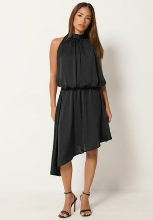 Femme portant une robe asymétrique noire sans manches avec un col haut et des talons hauts noirs à bride à la cheville, debout devant un fond clair.