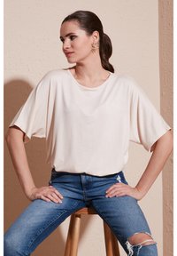Světlý béžový oversized tričko s krátkými rukávy, hladký povrch, kulatý výstřih. V kombinaci s modrými roztrhanými skinny džíny, sedící na dřevěné stoličce.