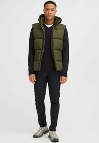 Mörkgrön vadderad väst med huva över en svart tröja, tillsammans med svarta byxor och beige-grå sneakers. Enkel design och funktionell passform.