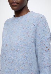 Gros plan d'une personne portant un pull texturé bleu clair avec des taches multicolores et un col rond sur fond uni.
