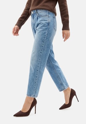 Persona che cammina indossando jeans dritti azzurri, scarpe con tacco marroni e un maglione marrone a maniche lunghe.