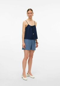 Navy blauw mouwloos topje met geplooide halslijn, gecombineerd met lichtblauwe shorts en witte blokhak schoenen. Eenvoudige, casual outfit.