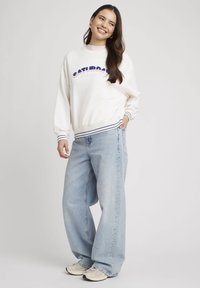 Sweatshirt blanc avec "SATURDAY SOCIAL CLUB" en texte bleu et rose, associé à un jean large bleu clair et des baskets beiges.
