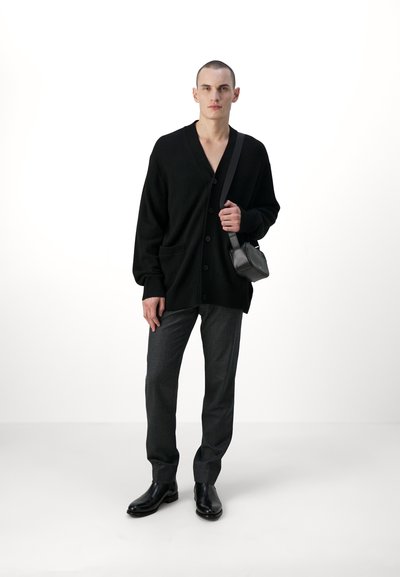 Cárdigan de punto negro con un escote en V profundo, cierre de botones y bolsillos frontales; combinado con pantalones gris oscuro y botas de cuero negras.