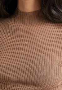 Gros plan sur une personne portant un pull à col roulé côtelé de couleur marron clair, avec de longs cheveux foncés visibles sur les bords.