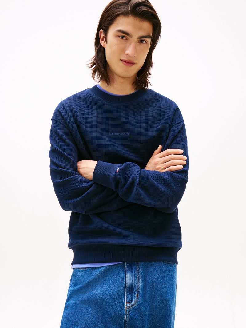 Sudadera de punto waffle azul marino con cuello redondo y puños acanalados, con un sutil logotipo en el pecho y superpuesta sobre una camisa azul.