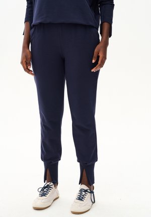 Osoba ubrana w granatowe spodnie dresowe z przednimi rozcięciami przy kostkach oraz białe sneakersy z granatowymi sznurowadłami, stojąca na białym tle.