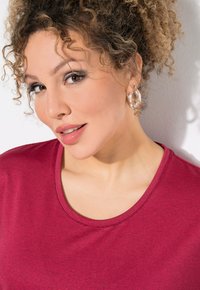 Donna con capelli ricci, che indossa una maglietta a righe rosse e orecchini a cerchio argentati, sorridendo leggermente, su uno sfondo di parete chiara.