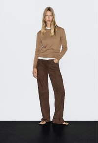 Brauner Strickpullover kombiniert mit braunen suède Schlaghosen; flache Sandalen sichtbar. Das Model steht vor einem neutralen Hintergrund.