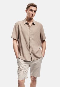 Lysbrun kortærmet skjorte med knapper i et struktureret stof, parret med beige shorts. Begge dele har en afslappet pasform og diskrete detaljer.