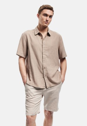 Chemise à manches courtes beige clair avec boutons, en tissu texturé, associée à un short beige. Les deux pièces sont d'une coupe décontractée et présentent des détails subtils.