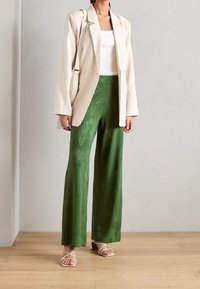 Béžové lněné sako přes bílý žebrovaný top bez rukávů, doplněné zelenými širokými kalhotami s potiskem a bílými sandály s pásky. Minimalistický design.