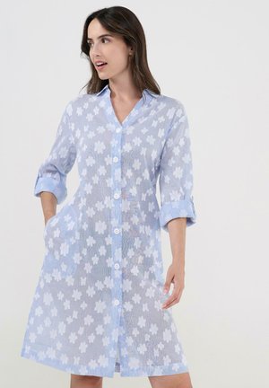 Mujer con un vestido azul claro abotonado con patrones florales blancos y mangas remangadas, de pie frente a un fondo blanco liso.