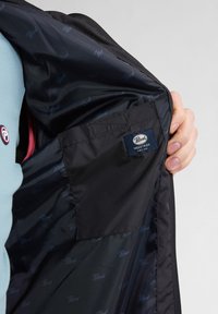 Interno della giacca nera con una texture liscia, etichetta blu scuro con la scritta "Petrol Industries" e uno sfondo rosa a contrasto.