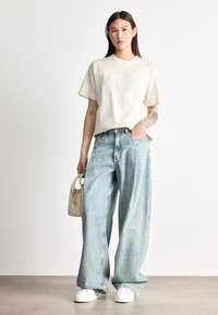 Κοντομάνικο λευκό μπλουζάκι με το λογότυπο "BOSS", σε συνδυασμό με ανοιχτό μπλε τζιν παντελόνι με φαρδιές μπάλες και λευκά αθλητικά παπούτσια. Το μοντέλο κρατά μια μικρή μπεζ τσάντα χειρός.