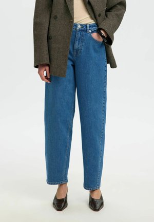 Personne portant un jean bleu taille haute, un blazer marron foncé, un haut beige et des chaussures foncées à bout pointu, debout avec une main dans la poche.