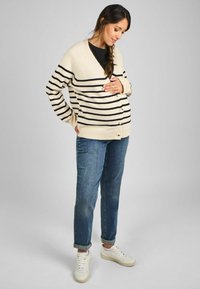 Cardigan beige con strisce nere, scollo a v, chiusura con bottoni, polsini a coste, abbinato a jeans blu e sneakers bianche.
