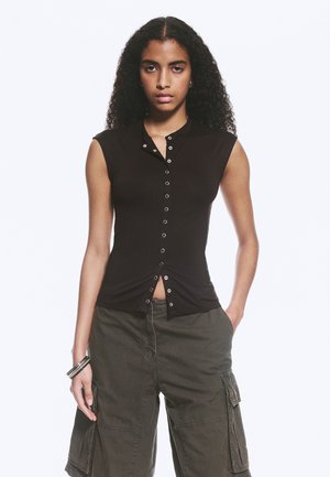 Jeune femme aux cheveux bouclés porte un haut noir sans manches boutonné et un short cargo sombre, debout avec une main dans la poche et une expression neutre.