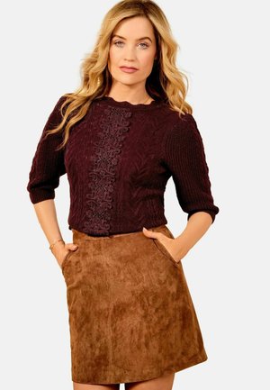 Frau in einem dunkelburgunderfarbenen strukturierten Strickpullover und einem braunen Wildleder-A-Linie-Rock mit den Händen in den Taschen.