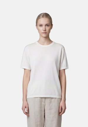Femme portant un t-shirt blanc uni à manches courtes et un pantalon beige, debout devant un fond clair uni, face à l'avant avec une expression neutre.