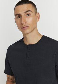 Tumši zils kokvilnas henley krekls ar apaļu kakla izgriezumu un pogu slēgtu trīsstūrveida apdari ar trim melniem pogām, strukturēta auduma un īsām piedurknēm.