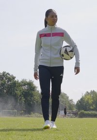 Adidas tepláková súprava v svetlo sivej farbe s ružovými akcentmi, spárovaná s tmavými nohavicami. Subjekt drží čierno-biely futbalový loptu na trávnatej ploche.