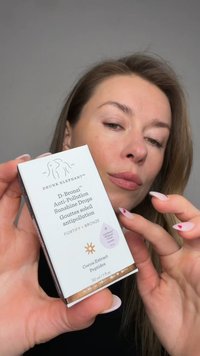Woman holding Drunk Elephant D-Bronzi Anti-Pollution Sunshine Drops skincare product near face, showcasing packaging text and logo.