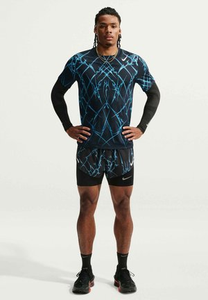 Homme athlétique debout, mains sur les hanches, portant une tenue de course noire avec des motifs abstraits bleus, des manchettes noires et des chaussures de sport.