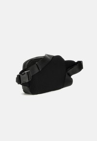 Sac noir avec un extérieur en maille texturée, doté d'une sangle noire lisse et d'une boucle. Design compact et courbé avec une ouverture zippée.