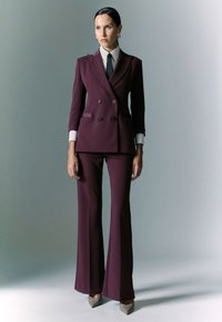 Completo su misura bordeaux scuro con giacca doppiopetto, camicia bianca, cravatta nera e pantaloni svasati, abbinato a scarpe grigie a punta.