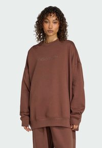 Hnědá oversized mikina vyrobená z měkkého materiálu, s kulatým výstřihem a vyšitým logem na prsou. Dlouhé rukávy s žebrovanými manžetami.